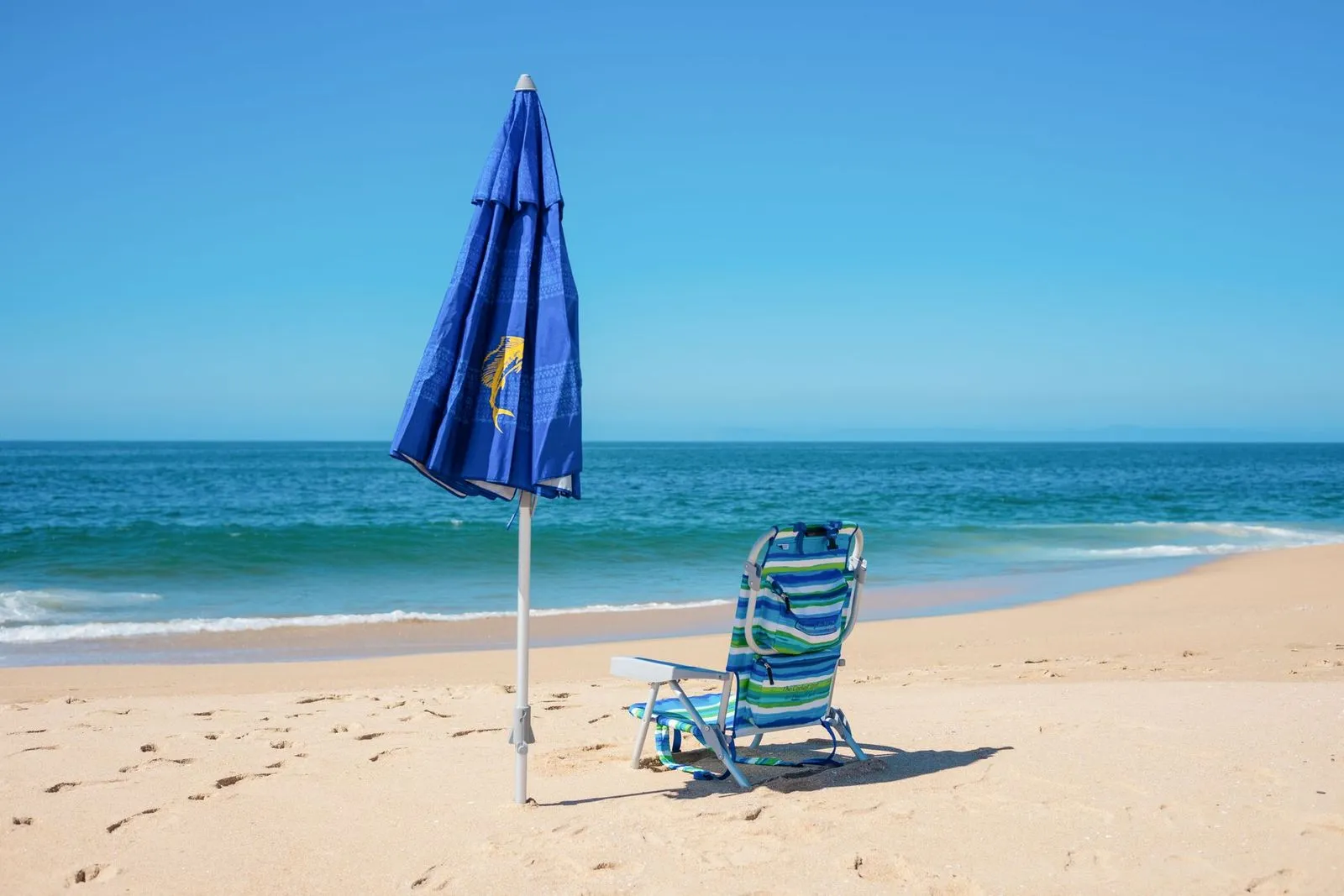 Guarda-sol azul fechado e cadeira de praia listrada sobre areia clara, de frente para o mar azul-turquesa em dia de sol.