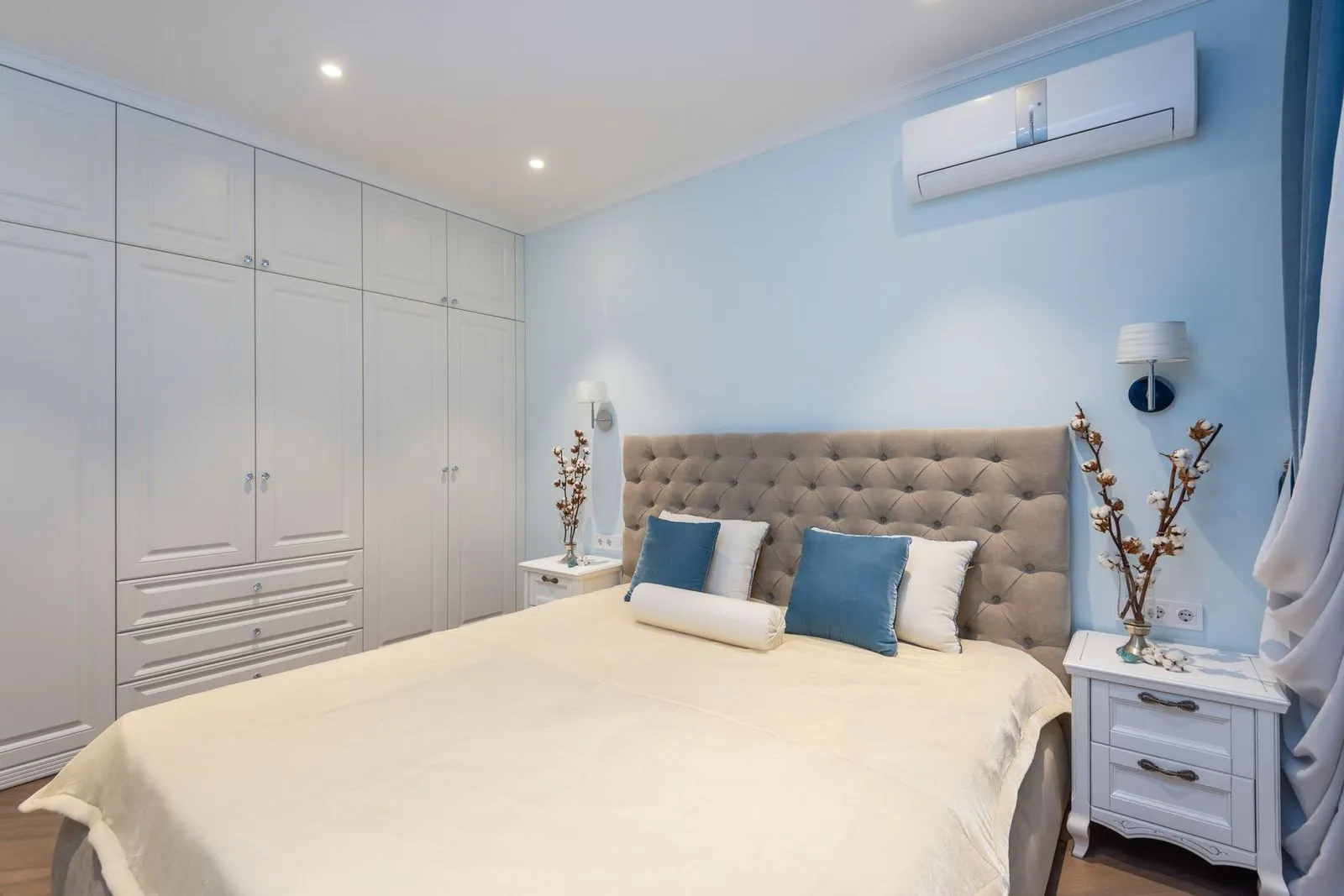 Quarto claro com parede azul-clara e um aparelho de ar-condicionado split branco instalado na parede, perto do teto, e cama com roupa de cama clara.