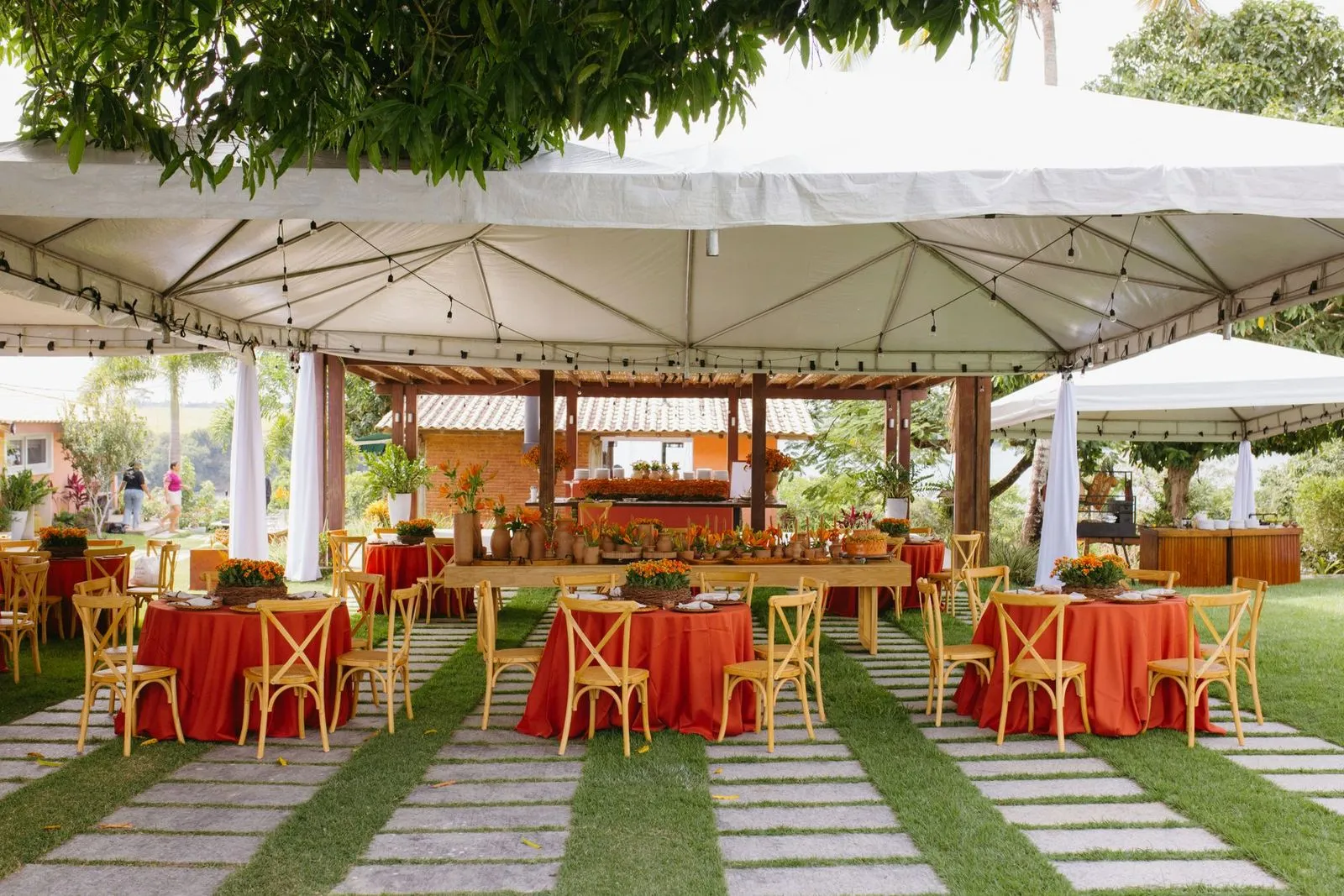 Montagem de festa ao ar livre sob um toldo branco amplo, com mesas de toalha vermelha, cadeiras de madeira e decoração laranja e vermelha no gramado.