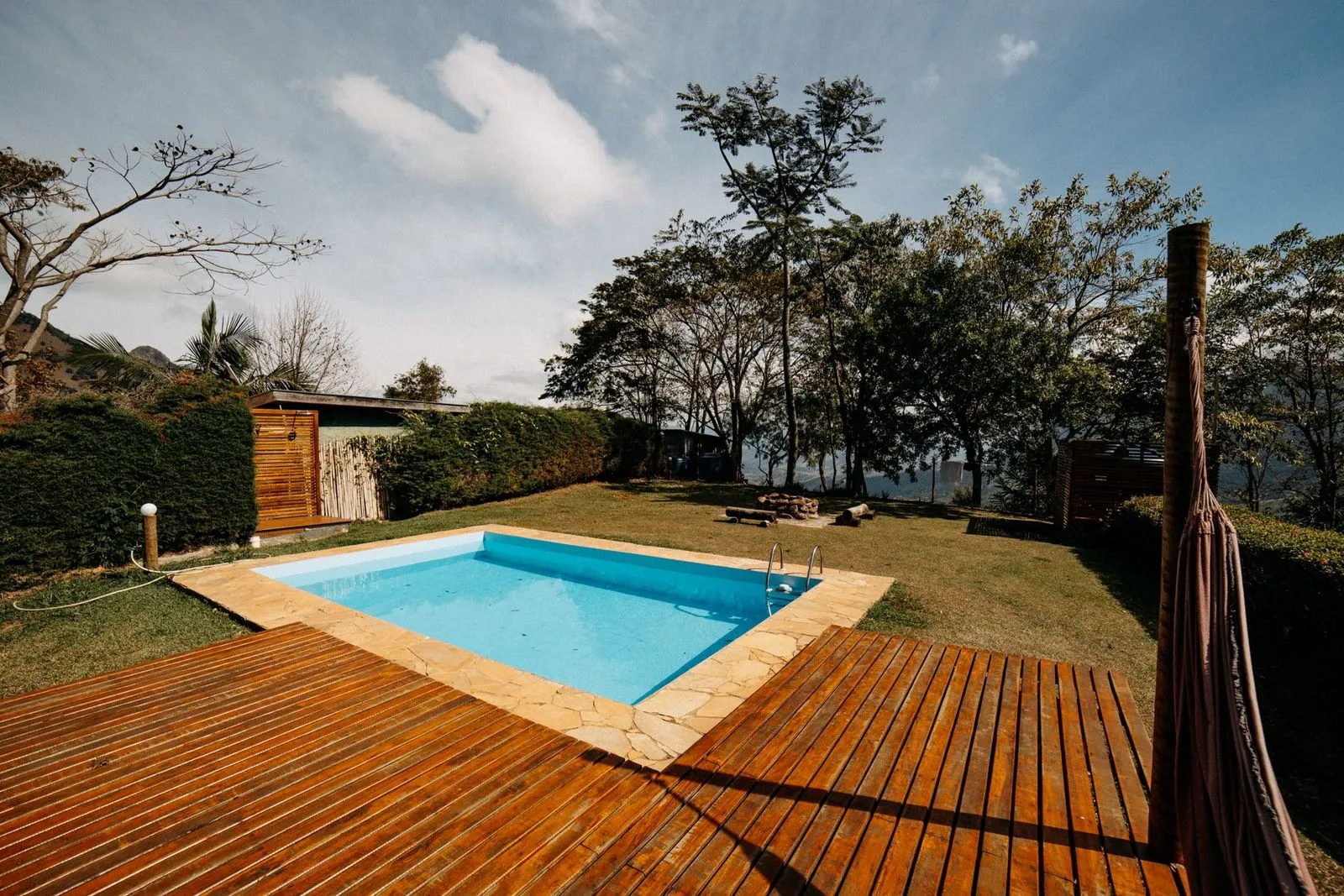 Piscina ao ar livre com deque de madeira e vegetação exuberante ao redor, sob céu claro.
