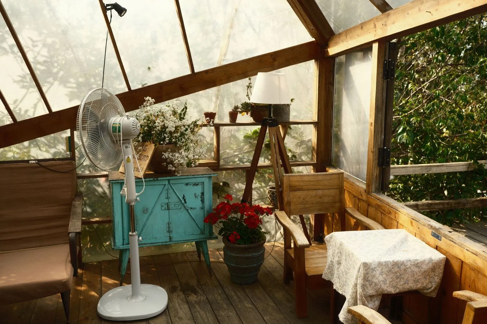 Ventilador de pé branco numa varanda de vidro ensolarada, com mesa de madeira coberta por toalha e armário azul-turquesa ao fundo.